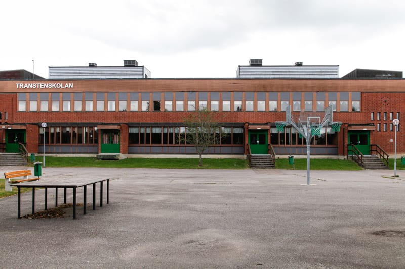 En fasad av en tegelbyggnad med namnet "TRANSTENSKOLAN" skrivet på den. Framför byggnaden syns asfalt, gräsmattor, träd, bänkar, en basketkorg med flera ringar och några soptunnor. Flera gröna dörrar och fönster är synliga på fasaden.