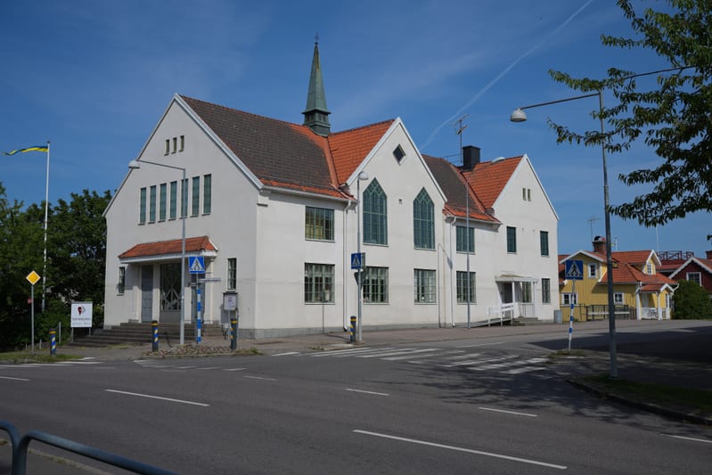 En gathörna med en stor vit byggnad med rött tak och en liten spira, samt flera mindre byggnader i bakgrunden. En väg och gångväg med zebratecknade övergångsställen syns i förgrunden, och himlen är blå med ett flygplansspår.