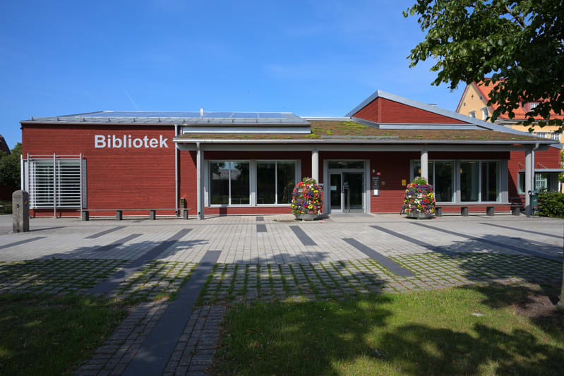 En röd byggnad med texten "Bibliotek" på fasaden sett diagonalt framifrån. Taket är delvis täckt av sedum och solpaneler. Framför byggnaden finns ett stenlagt område med inslag av gräs och två blomsterurnor. Höger om byggnaden syns trädtoppar och en