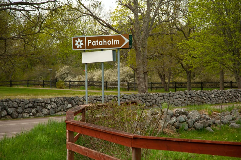 En vägskylt med texten "Pataholm" och en vit blomsymbol står vid sidan av en väg. I förgrunden syns en röd träspång och gräs. En stenmur löper längs vägen, och i bakgrunden finns gröna träd och något som ser ut som blommande buskar.
