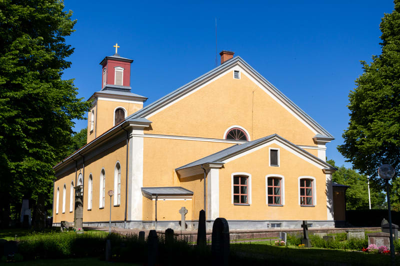 En gul kyrkobyggnad med gråa tak och en rödspetsig klockstapel som kröns av ett gyllene kors, under en klarblå himmel. Runt kyrkan finns gröna träd och en kyrkogård med gravstenar och ett kors. Solen lyser på byggnaden.