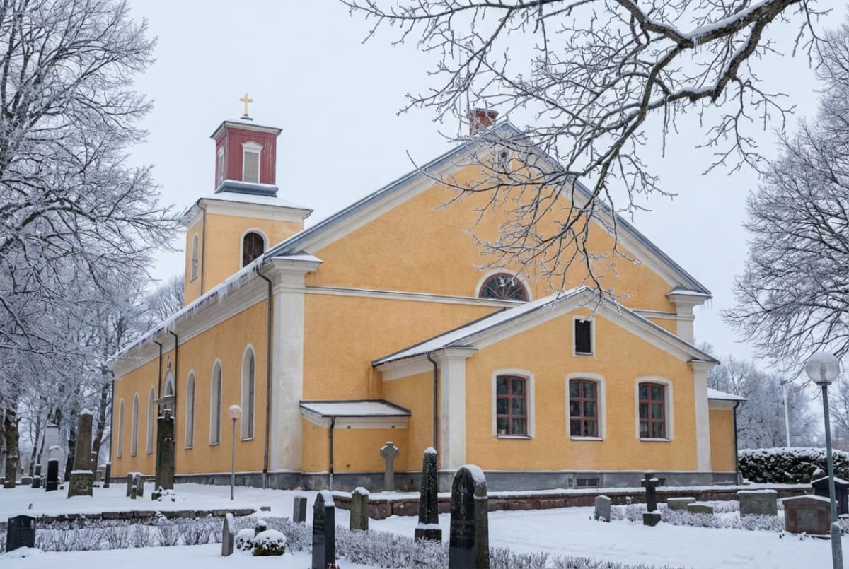 En gul kyrka med rött tak och ett guldfärgat kors på toppen, delvis täckt med snö och omgiven av snötäckta träd och gravstenar. Istappar hänger från taket.