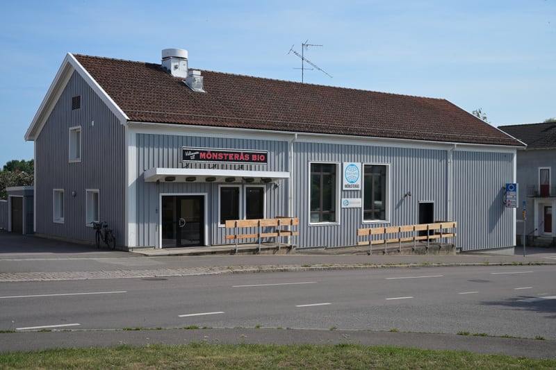 En enplansbyggnad med grå träfasad och brunt tegeltak står bredvid en asfalterad väg. På fasaden finns skyltar för "MÖNSTERÅS BIO" och en logotyp för "MONTERAS". Framför byggnaden finns träbänkar och en cykel. Vägen har vita streckmarkeringar.