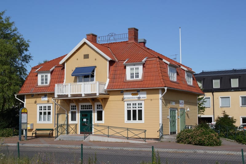 En gul byggnad med rött tak och vita detaljer står under en klarblå himmel. Utanför byggnaden finns en bänk, en busskylt, en ramp och buskage. En skylt med texten "INFORMATION & FÖRSÄLJNING" syns på fasaden.