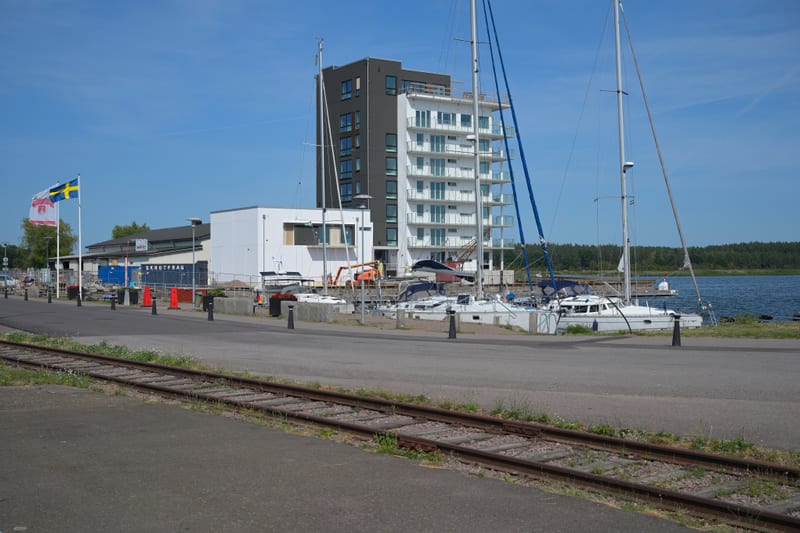 En modern byggnad under uppförande med en fasad i svart och vit, med balkonger mot vattnet. Flera segelbåtar ligger förtöjda i vattnet intill en kaj. Bredvid byggnaden syns anläggningsarbete. I förgrunden syns järnvägsräls, och i bakgrunden en svensk