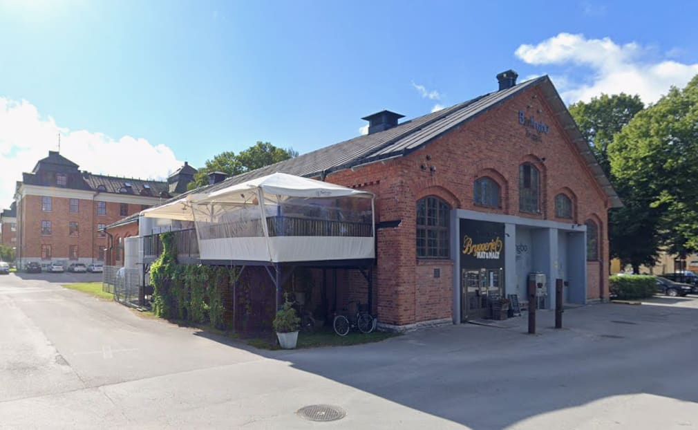 En stor röd tegelbyggnad med ett svart tak, där en uteservering med vitt tälttak är fäst på sidan. Ett skyltfönster med texten "Bryggeriet Mat & Malt" syns på fasaden. I bakgrunden finns en annan tegelbyggnad och träd. Parkerade bilar syns i förgrund