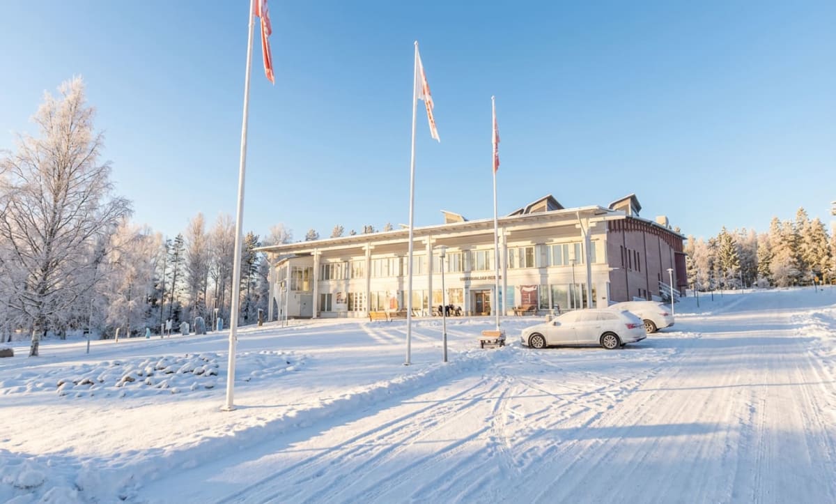 En exteriör vy av en byggnad på en snötäckt dag. Byggnaden är vit med stora fönster och en mörkare sektion på höger sida. Flera flaggstänger med flaggor står framför byggnaden. Två bilar är parkerade i snön till höger om byggnaden, och det finns träd