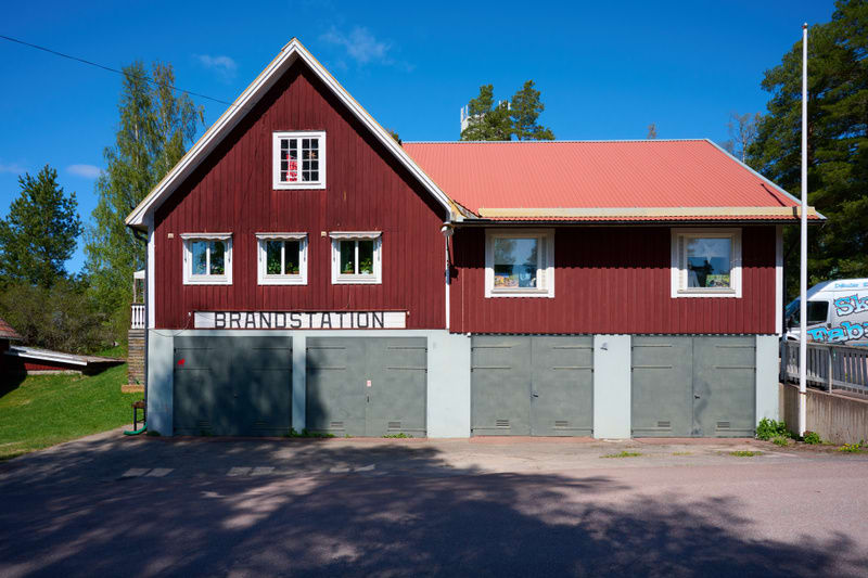 En röd träbyggnad med vita fönsterkarmar och en röd takpannor under en klarblå himmel. På fasaden står det "BRANDSTATION" och nedanför finns flera grå garageportar. Asfalt med skuggor framför byggnaden och grönska i bakgrunden.