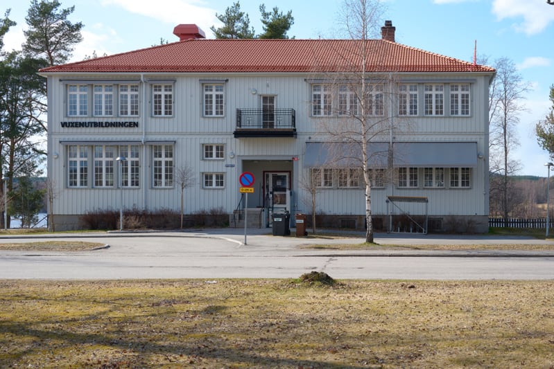 En ljus byggnad med rött tak och skylten "VUXENUTBILDNINGEN" på fasaden. Framför byggnaden finns en asfalterad väg och en gräsmatta. I bakgrunden syns skog och en sjö. Träd utan löv står nära byggnaden och på gräsmattan.