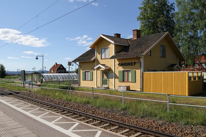 En gul trähusbyggnad med texten "GAGNEF" på fasaden står bredvid ett järnvägsspår och en perrong under en klarblå himmel med några moln. Till vänster om byggnaden finns en cykelparkering under tak samt gatubelysning, och i bakgrunden syns skog.