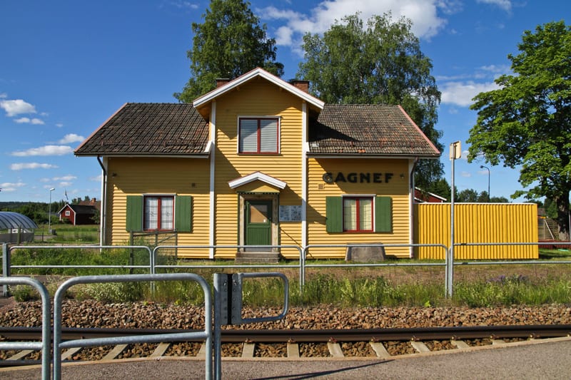 En gul byggnad med texten "GAGNEF" på väggen står vid en järnvägsräls under en blå himmel med vita moln. Byggnaden har bruna takpannor och gröna fönsterluckor. Framför rälsen finns en metallbarriär och gräs.