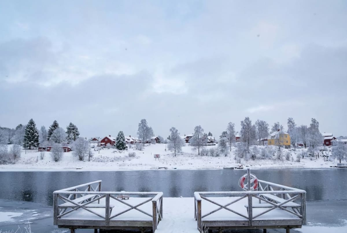 En vinterbild över en vattensamling med snötäckta byggnader och träd på motsatt sida. I förgrunden syns två snötäckta träbryggor. En livboj med röda och vita sektioner hänger på räcket till den högra bryggan. Himmeln är molnig och ljusgrå.