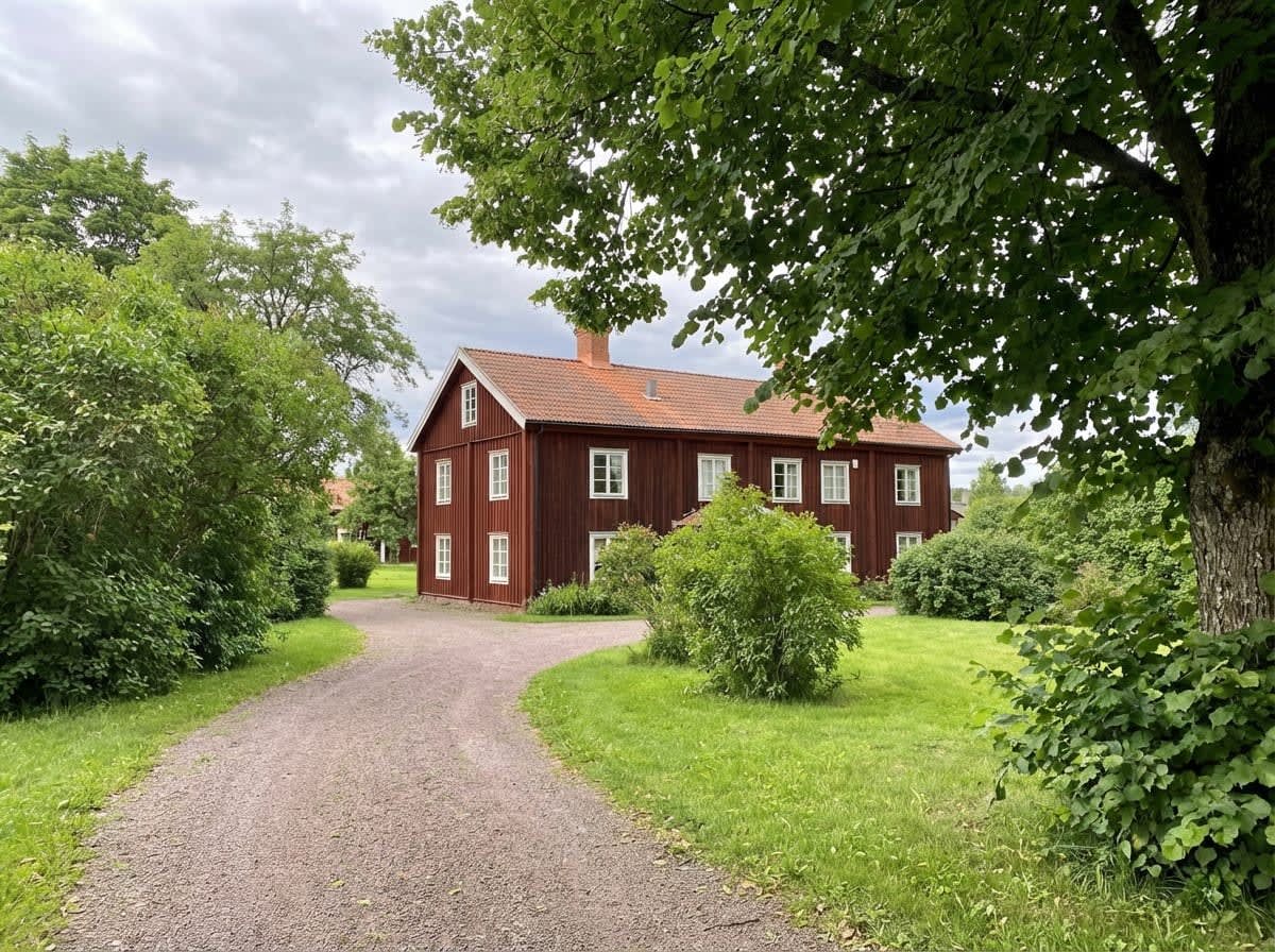 En stor, röd träbyggnad med vita fönster och rött tegeltak, syns bakom en grusväg som svänger. Byggnaden omges av gröna träd och buskar, och en gräsmatta i förgrunden. Himlen är mulen.
