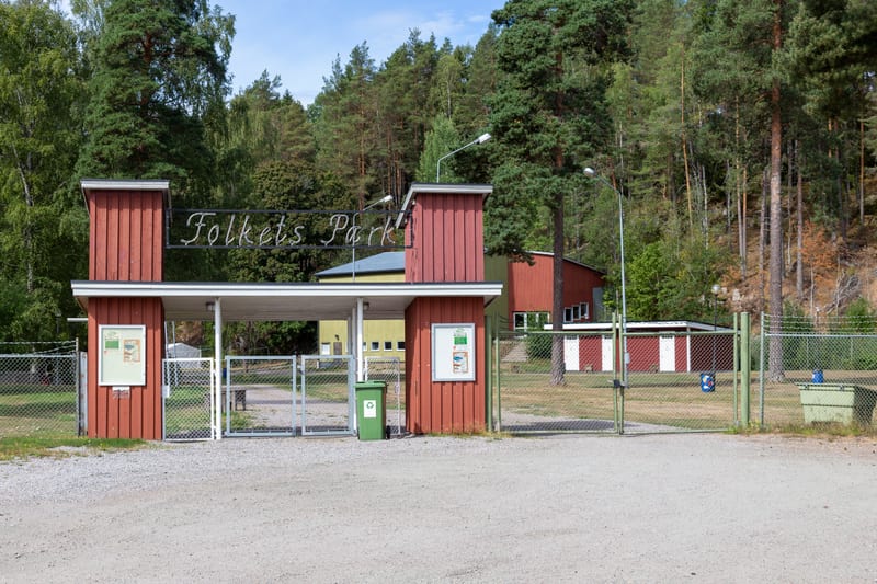 Bild på entrén till "Folkets Park" som består av två röda pelare med en skylt över. Framför finns en grusplan och bakom syns byggnader, en metallgrind, en grön soptunna och skogsklädda kullar under en molnig himmel.