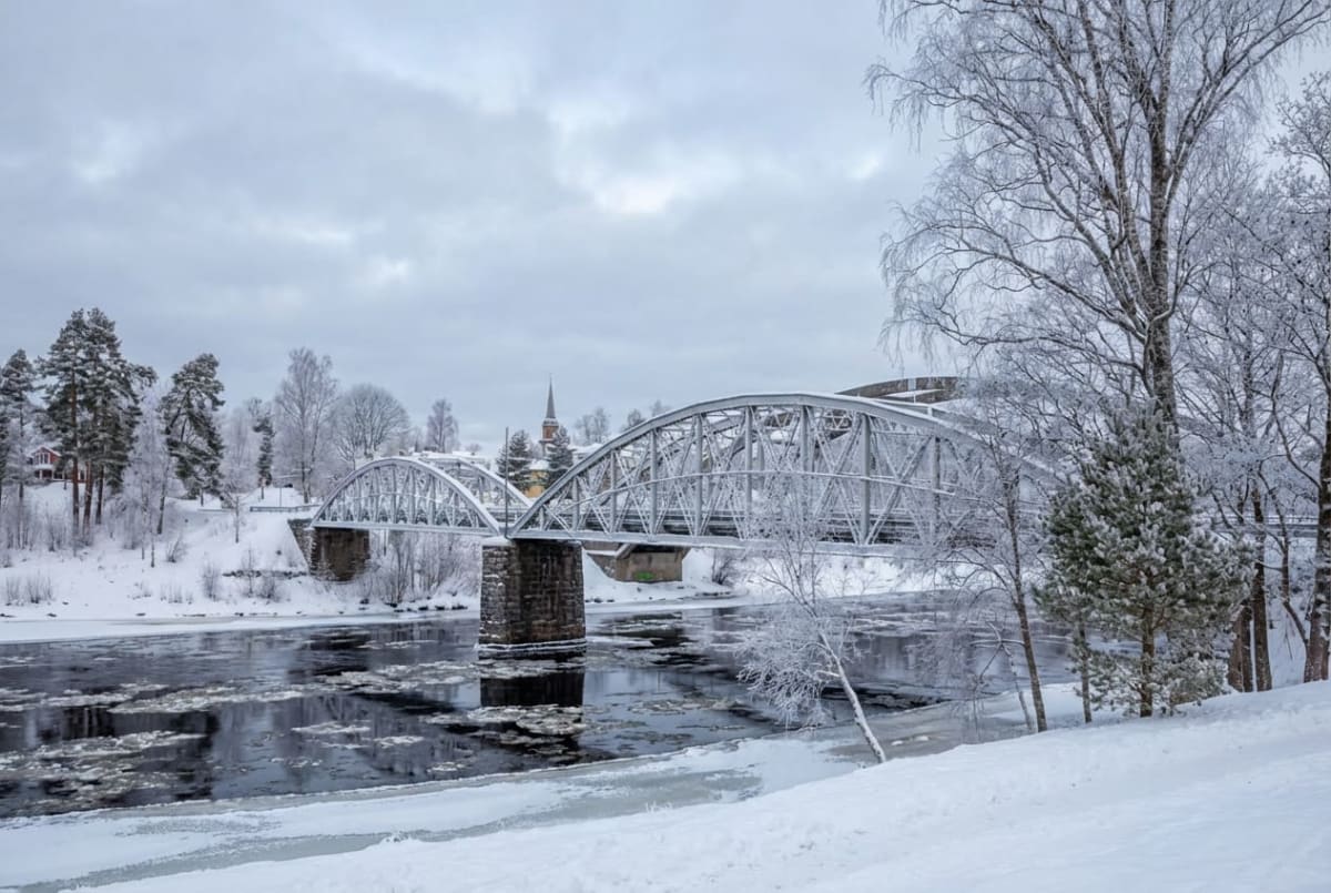 En vinterbild från ett snöigt landskap där en flod med isflak flyter under en metallbro med valv. Träden längs floden och i bakgrunden är täckta av snö och frost. En byggnad med ett spetsigt torn är synlig i fjärran. Himlen är mulen.