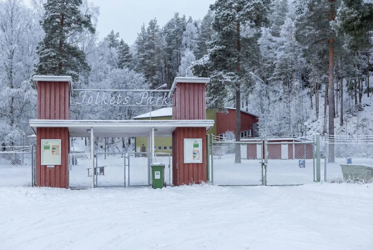 En entré till "Folkets Park" täckt i snö. Entrén består av två röda byggnader med ett tak mellan dem, och en metallgrind. Skylten "Folkets Park" är monterad ovanför entrén. I bakgrunden syns snötäckta träd och byggnader.