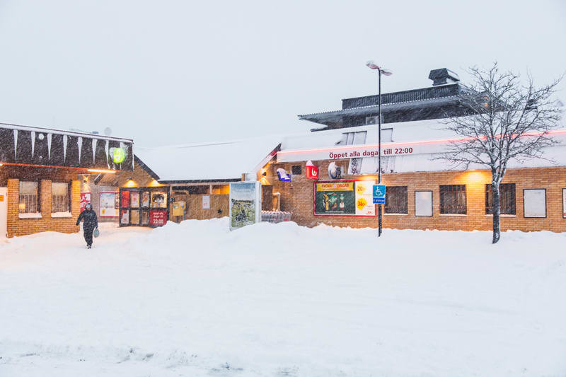 En person går framför en tegelbyggnad under snöfall på kvällen. Byggnaden har ett snötäckt tak med istappar och skyltar med texten "Öppet alla dagar till 22:00". Stor mängd snö täcker marken och en bar trädkrona syns till höger.