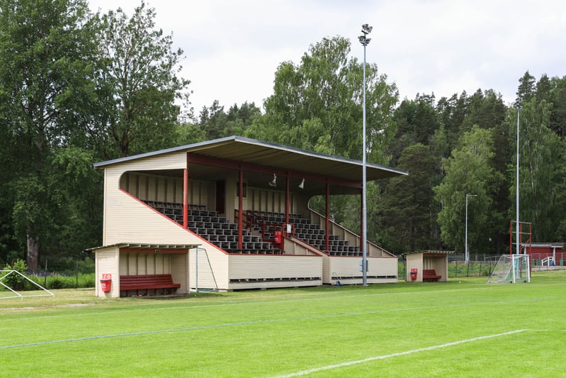 En bild av en tom fotbollsplan med en träläktare i bakgrunden. Bakom läktaren syns träd och en gråmulen himmel. På fotbollsplanen finns vita linjer, två bänkar och ett litet mål. Två höga belysningsstolpar står vid läktaren.