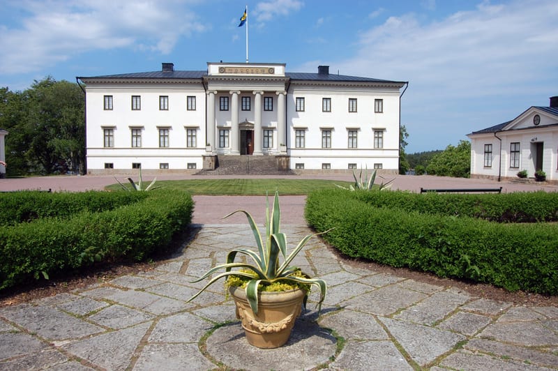 En stor vit byggnad med pelare vid ingången och en svensk flagga på taket. Framför byggnaden finns en trädgård med en gång, buskar och en stor kruka med en växt. Till höger finns en mindre, liknande byggnad.