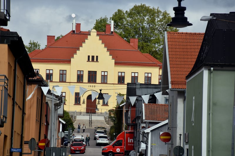 En gata i en stad med byggnader på båda sidor och en större gul byggnad med rött tak i bakgrunden. Ett rött fordon och flera personer syns på gatan. Vita vimplar hänger över gatan.