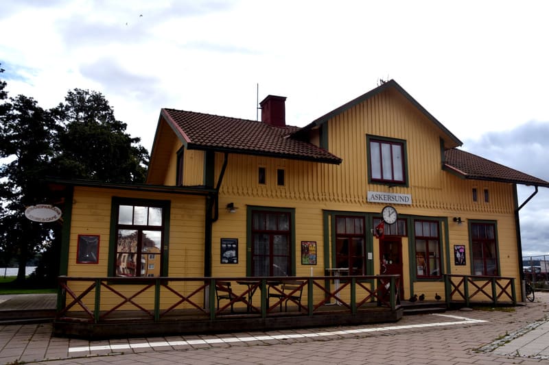 En gulmålad träbyggnad med rött tak och mörkgröna detaljer, under en molnig himmel. Byggnaden har ordet "ASKERSUND" skrivet på framsidan under ett fönster och en klocka. På marken framför byggnaden finns ett trästaket och stenplattor. En skylt med "S