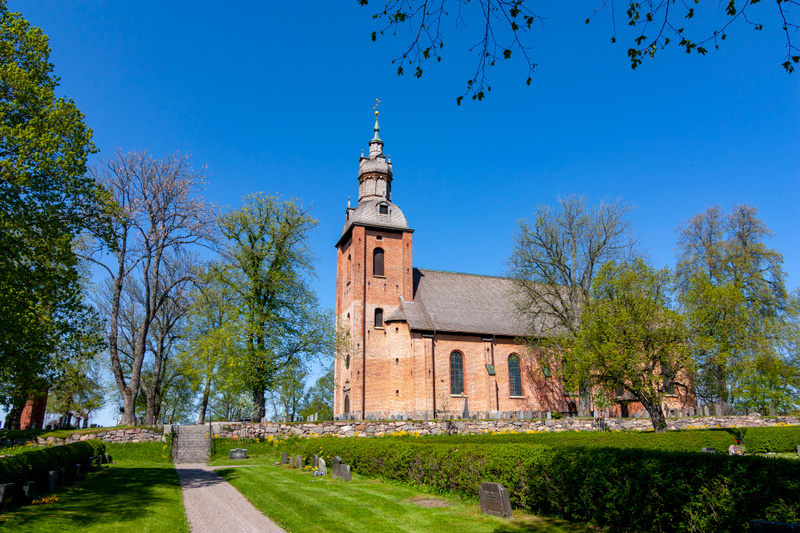 En röd tegelkyrka med en tornspira under en klarblå himmel. Kyrkan är omgiven av gröna gräsytor, träd och en kyrkogård med gravstenar. En grusgång leder fram till en stentrappa.