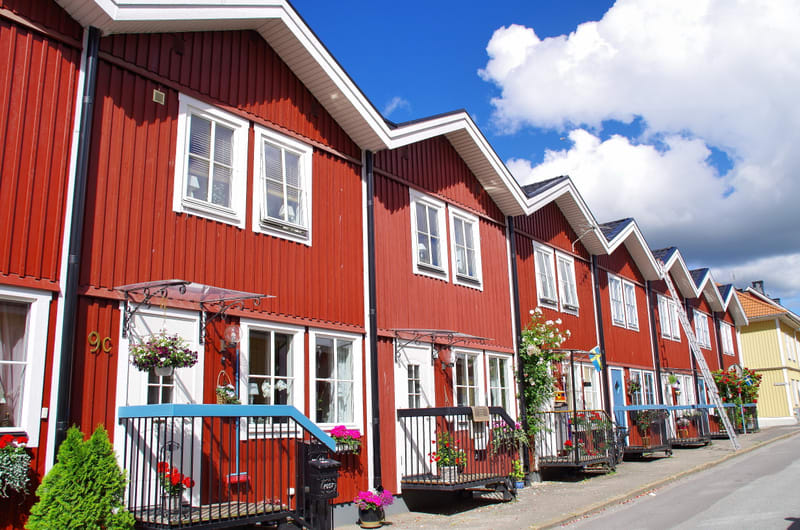 En rad husfasader målade i rött med vita fönster och dörrkarmar syns under en blå himmel med vita moln. Husen har vita snedtak och ut mot gatan finns små verandor med räcken. Blommor syns på verandorna och en stege står lutad mot ett av husen.