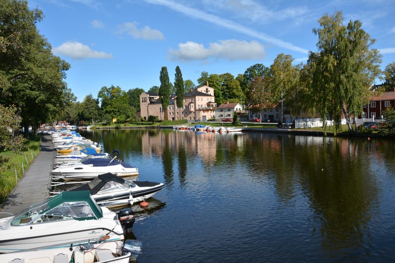 En översiktsbild av en kaj med flera båtar förtöjda längs en träbrygga, vid en vattenkanal. På andra sidan kanalen syns byggnader i olika färger samt träd med grönskande lövverk under en blå himmel med några moln. Båtarnas reflektioner syns i vattnet