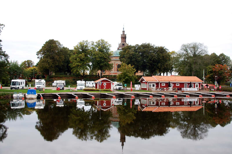 En bild som visar en småbåtshamn eller kaj med flera förtöjda båtar. I bakgrunden syns husbilar parkerade på ett grönområde, röda byggnader med vita knutar, träd och en kyrkobyggnad med en tornspira i fjärran. Vattnet reflekterar omgivningen.