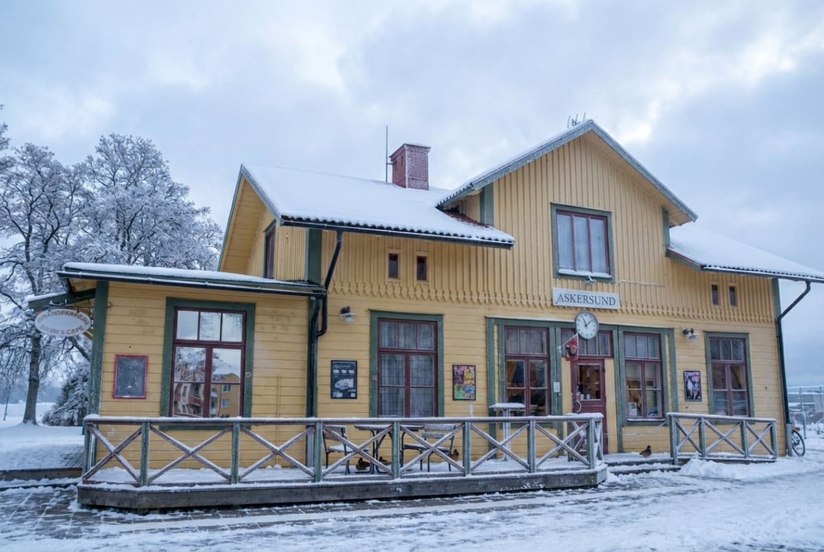 En gul byggnad med gröna och röda detaljer, täckt av snö, under en mulen vinterhimmel. En skylt med texten "ASKERSUND" syns ovanför ingången samt en annan skylt med texten "POLISENS CAFE". Byggnaden har en altan med staket och möbler, även de med snö