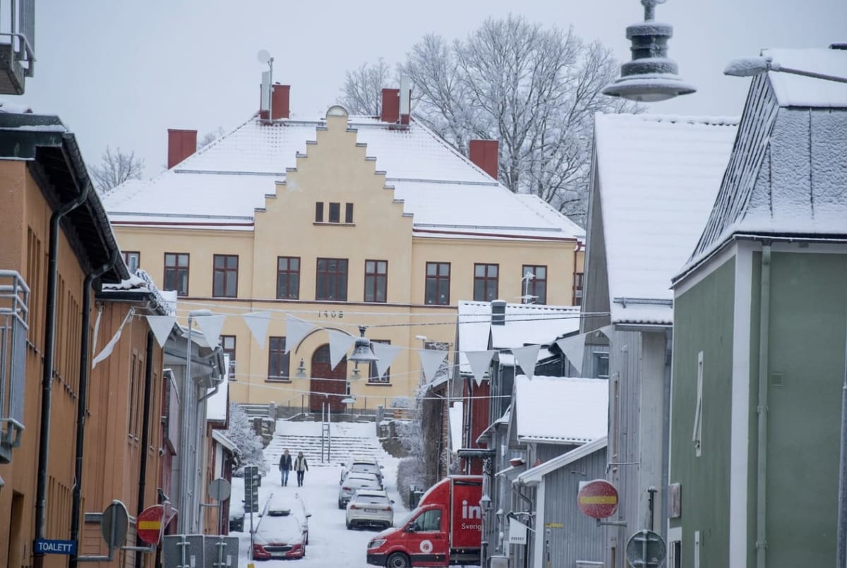 En gata i en stad täckt med snö. Byggnader med snö på taken kantar gatan, och i slutet av den snöiga gatan syns ett större ljust gult hus med röda tak och trappor. Flera bilar står parkerade längs gatan, som också är snötäckt. Ett par människor går u