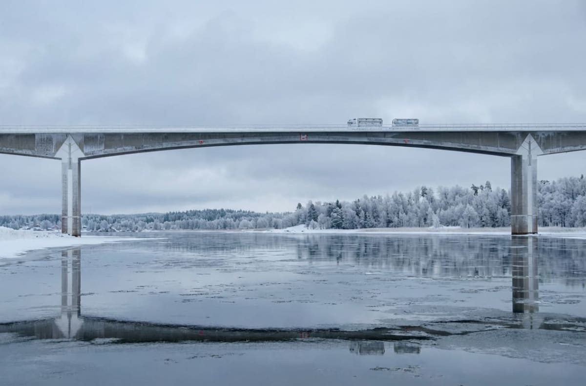 En betongbro sträcker sig över en vattensamling med isflak. På bron kör två lastbilar. Landskapet runt vattnet är täckt av snö och frostiga träd under en gråmulen himmel. Bron reflekteras i vattnet.