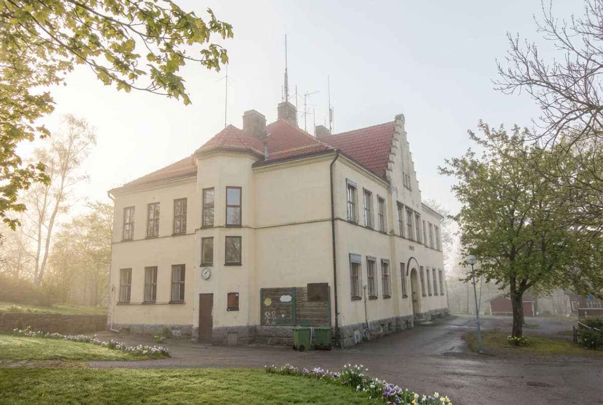 En gul byggnad med rött tak och prydnadsgavlar står i förgrunden, omgiven av gräsmatta, blommor och några träd. Morgondimma skymmer bakgrunden. Två gröna soptunnor syns bredvid byggnaden.