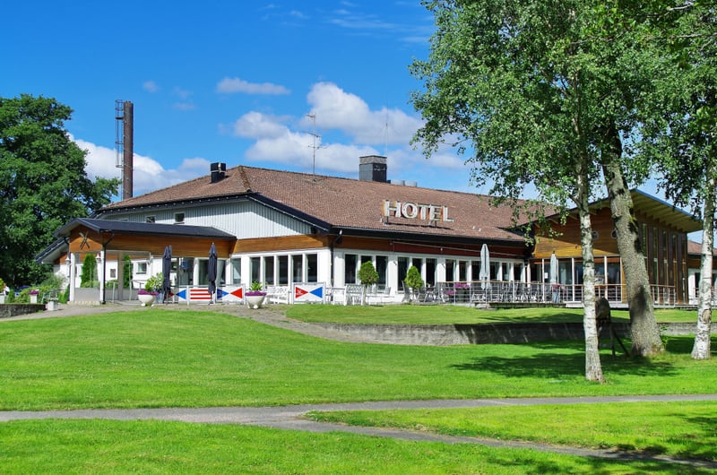 En stor byggnad med en tegeltäckt takskorsten och texten "HOTEL" på taket, omgiven av grön gräsmatta och flera lövträd under en blå himmel med vita moln. Framför hotellet finns en terrass med bord och parasoller samt flaggliknande dekorationer.