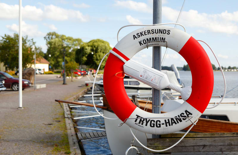 En röd och vit livboj med texten "ASKERSUNDS KOMMUN" och "TRYGG-HANSA" hänger på en stolpe vid en kajkant. I bakgrunden syns ett vattendrag, båtar, en parkeringsplats med bilar, träd och byggnader under en blå himmel med vita moln.