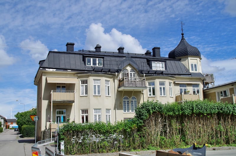 En stor beige byggnad med svart tak och flera fönster. Delar av taket är välvda, varav en toppas med en väderflöjel. Framför byggnaden finns en klippt häck och på marken står vad som ser ut som en trädgårdsvagn. Till vänster syns en gata med fler hus