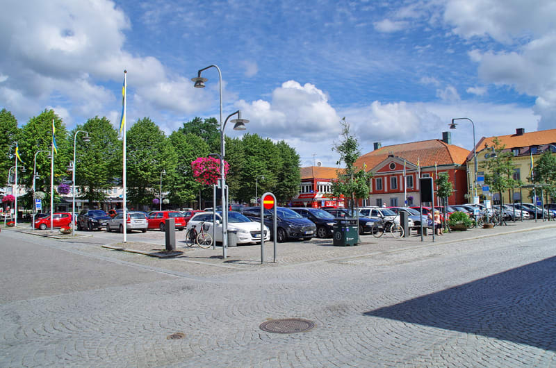 En utomhusbild som visar en gatuparkering med flera parkerade bilar, cyklar och stativ med flaggor i gult och blått. I bakgrunden syns flera byggnader och träd. Gatubeläggningen är av kullersten och himlen är molnig. Hängande blomlådor med rosa blomm