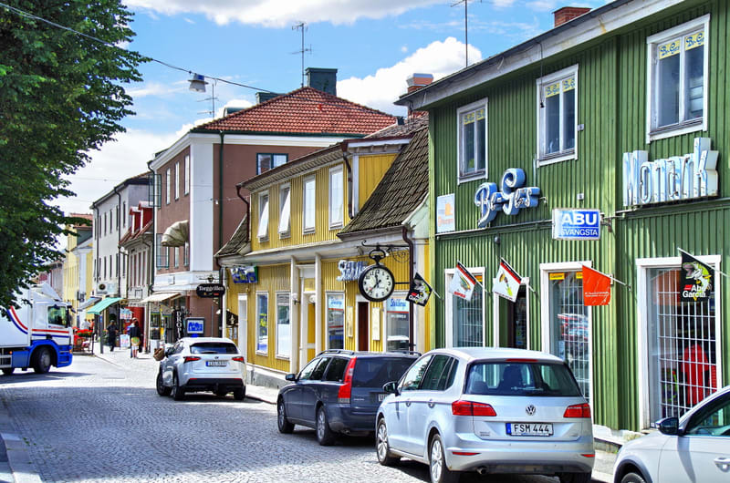 En gata med gatsten belagd med parkering på den högra sidan, med ett flertal bilar stående. Längs gatan kantar flertalet byggnader med butiker på bottenvåningen. Byggnaderna är byggda i olika stilar och har olika färger.