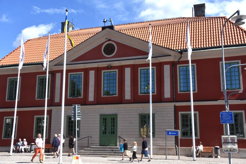 En röd byggnad med tegelrött tak och gröna fönsterramar under en blå himmel. Framför byggnaden står flaggstänger, varav en med svensk flagga. Människor syns utanför byggnaden, varav några sitter på bänkar. En skylt för "P-skivor" är också synlig.