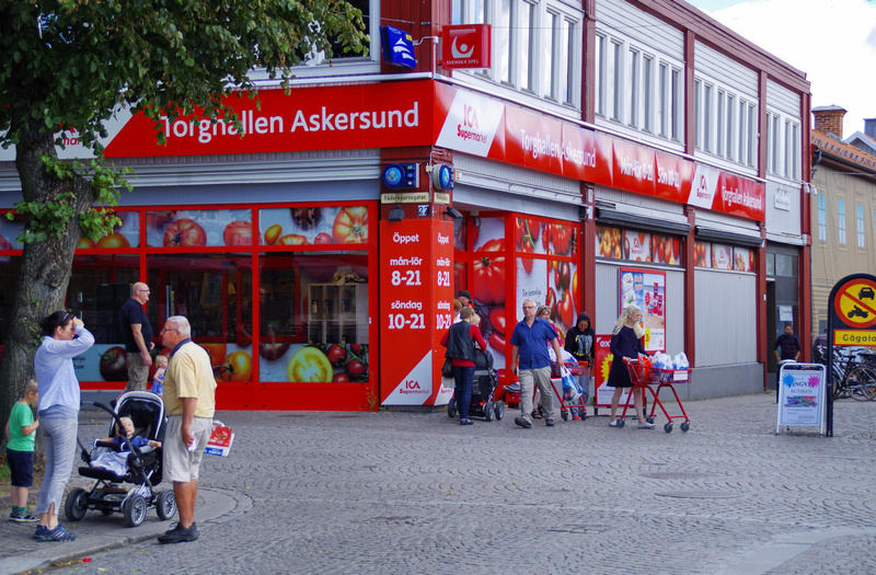 En gathörna med en ICA Supermarket, Torphallen Askersund, med stora skyltfönster. Flera personer står och går utanför butiken. En person med barnvagn och en ståendes bredvid. Butiken har röda skyltar och öppettider synliga. Fönstren visar bilder av g