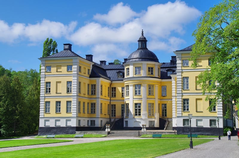 En stor, gul byggnad med många fönster och ett mörkt tak. Byggnaden har en kupol i mitten och trappor som leder upp till ingången. Framför byggnaden finns en gräsmatta med grusgångar, och träd omger området. En person syns gå på en grusgång till höge