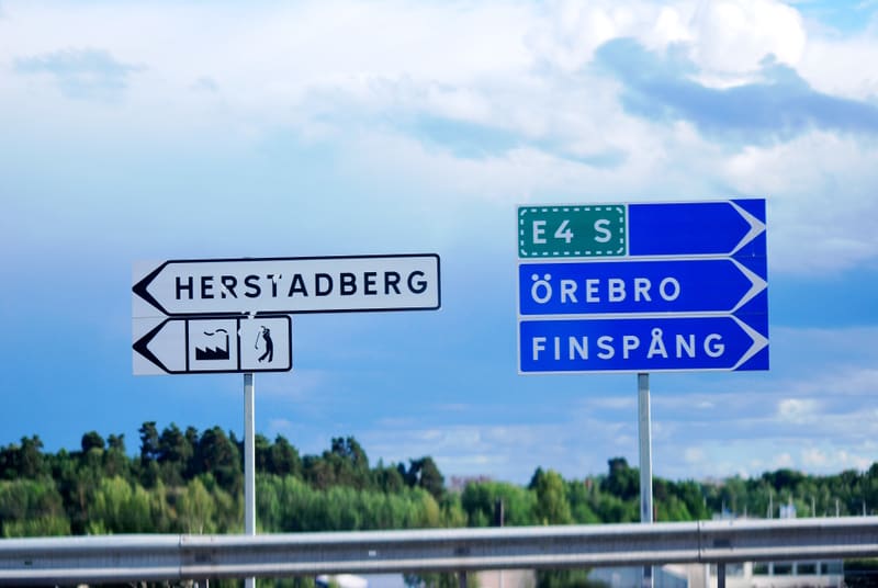 En vägskylt med destinationerna Herstadberg, Örebro och Finspång under en molnig himmel. Under Herstadberg finns symboler för en fabrik och en golfbana. Några gröna träd syns i bakgrunden.