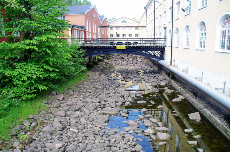 En bild av en grönskande kanal med stenar på botten, kantad av byggnader i olika färger och en bro. Vissa delar av kanalen har vatten, andra är torra.