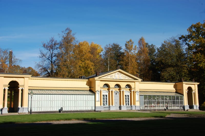 En gul byggnad med pelare och stora fönsterpartier syns i mitten av bilden. Byggnaden är omgiven av grönt gräs. I bakgrunden finns träd med gulaktiga löv och en blå himmel. Till vänster syns en skorsten.