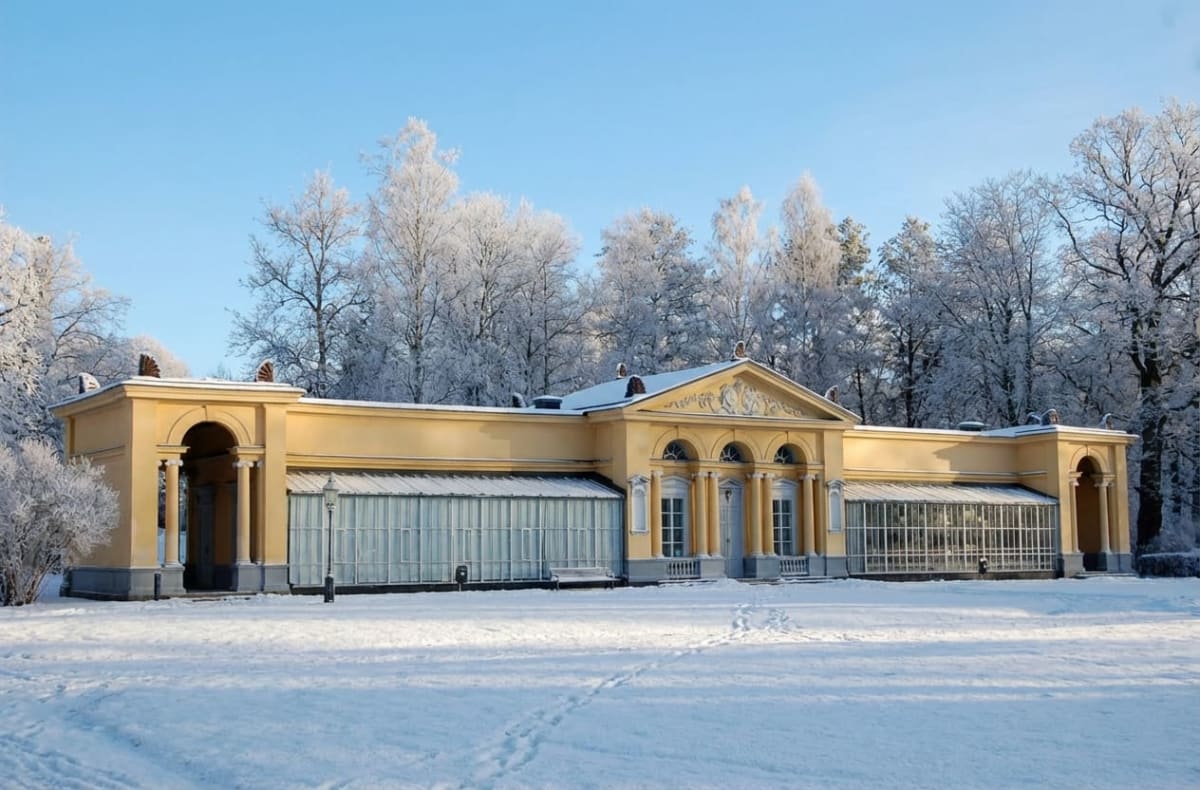En gul byggnad med stora fönster och pelare står mot en klarblå himmel. Mark och träd i bakgrunden är täckta av snö och frost. Fotspår syns i den orörda snön framför byggnaden.