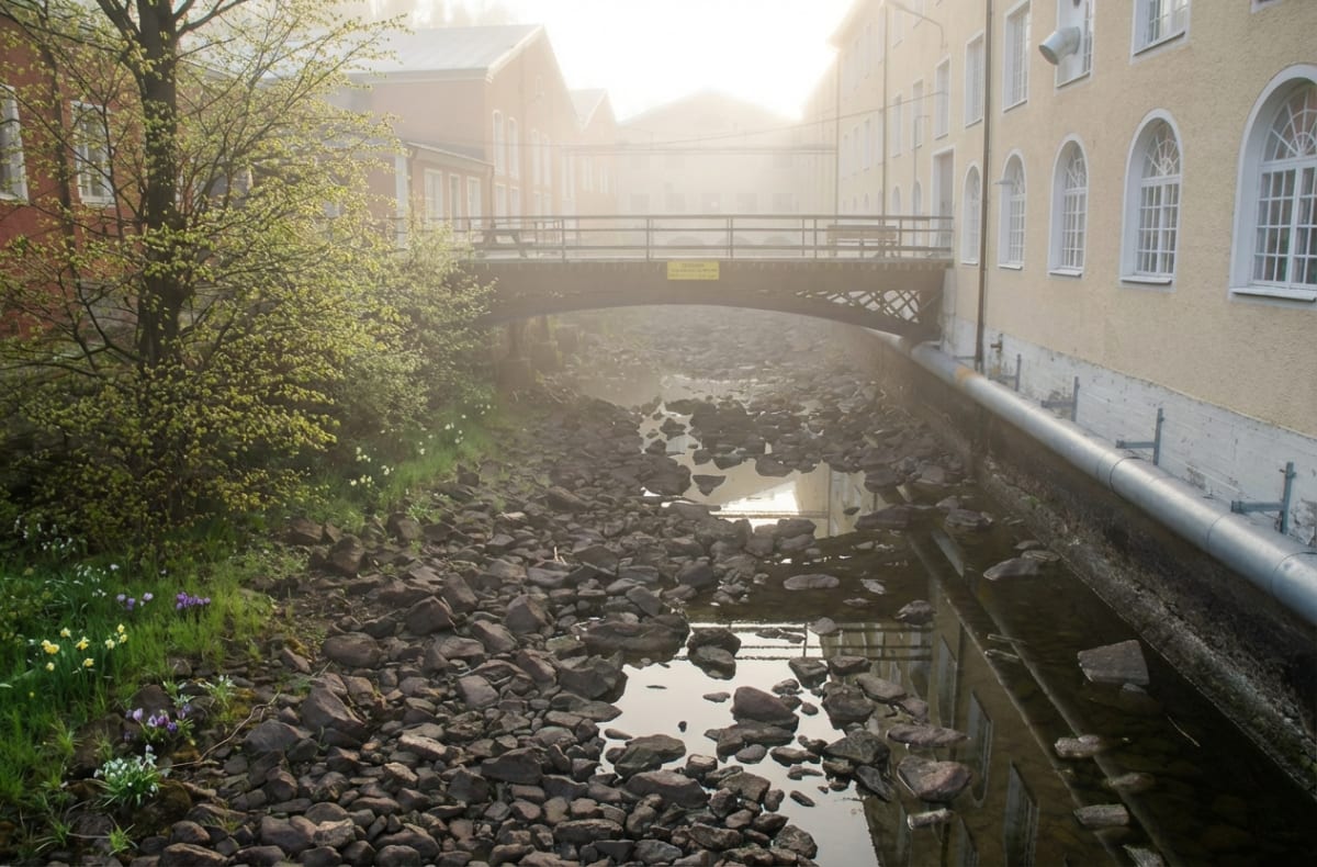 En bro sträcker sig över en stenfylld kanal med delvis stillastående vatten. På båda sidor av kanalen syns byggnader, varav en röd till vänster och en gul till höger. Båda har fönster. Vegetationen på vänster sida visar gräs och buskar med gröna knop