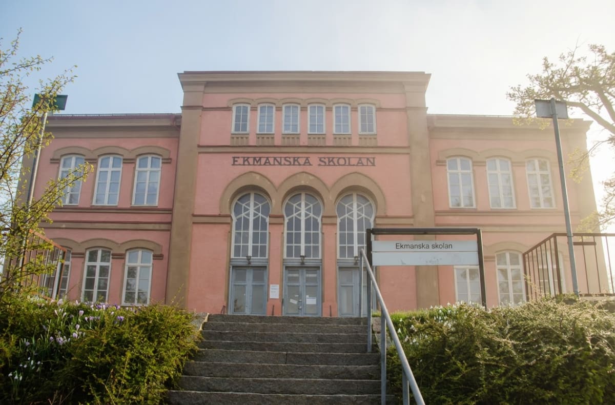 En rosa byggnad med texten "EKMANSKA SKOLAN" över tre stora fönster/dörrar med valv. Man ser en trappa som leder upp till ingången och buskar med vårblommor på sidorna. En skylt med texten "Ekmanska skolan" syns i bildens nedre högra del.