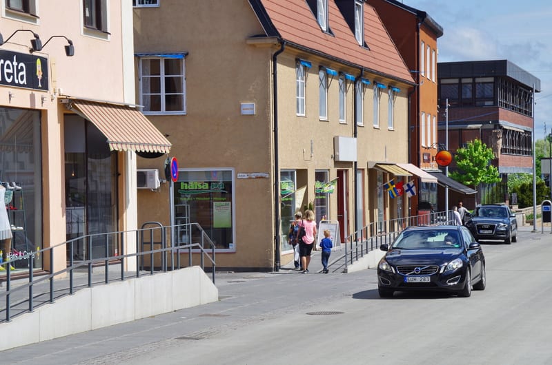 En gata framför en rad byggnader. En svart Volvo V60 färdas på körbanan i förgrunden, följt av en mörk SUV. Vid trottoaren syns flera personer, inklusive barn, utanför butiker med flaggor och markiser. Husfasaderna är beige och rosa, med ett tak i te