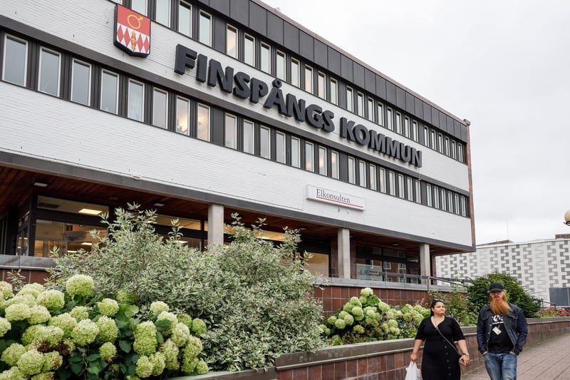 En bild som visar Finspångs kommunhus från en vinkel som fångar den vita tegelfasaden med skylten "FINSPÅNGS KOMMUN" i svart text. Framför byggnaden finns gröna buskage med ljusa blommor. Två personer går på en gångväg i förgrunden.