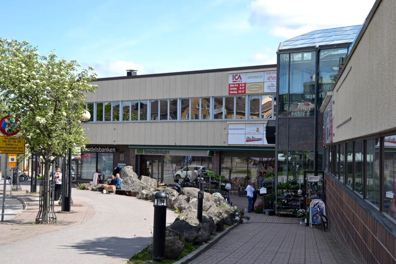 En utomhusbild som visar en gatuvy med flera byggnader och människor. I förgrunden syns en blommande buske och en gatuskylt. Till höger finns en stenlabyrint och en försäljningsplats för blommor. Byggnaderna har skyltar för butiker som ICA, Apotek oc